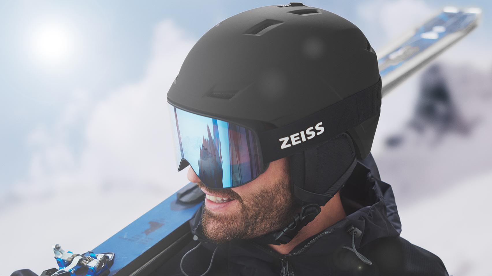Homem a usar capacete preto e máscara ZEISS para a neve com lente azul espelhada, a transportar os esquis sobre o ombro direito.